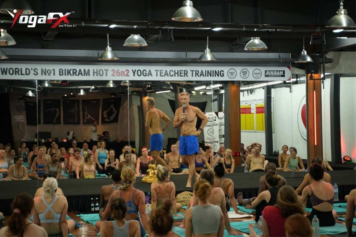 Teacher leading a Bikram Hot 26&2 class with a microphone at YogaFX Studio in Seminyak, with students seated facing the mirror