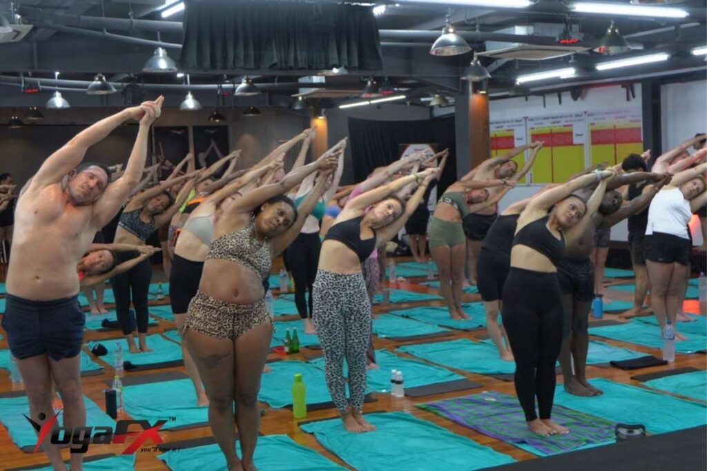 Students in a standing side bend in a crowded hot yoga class, with water bottles beside their towels