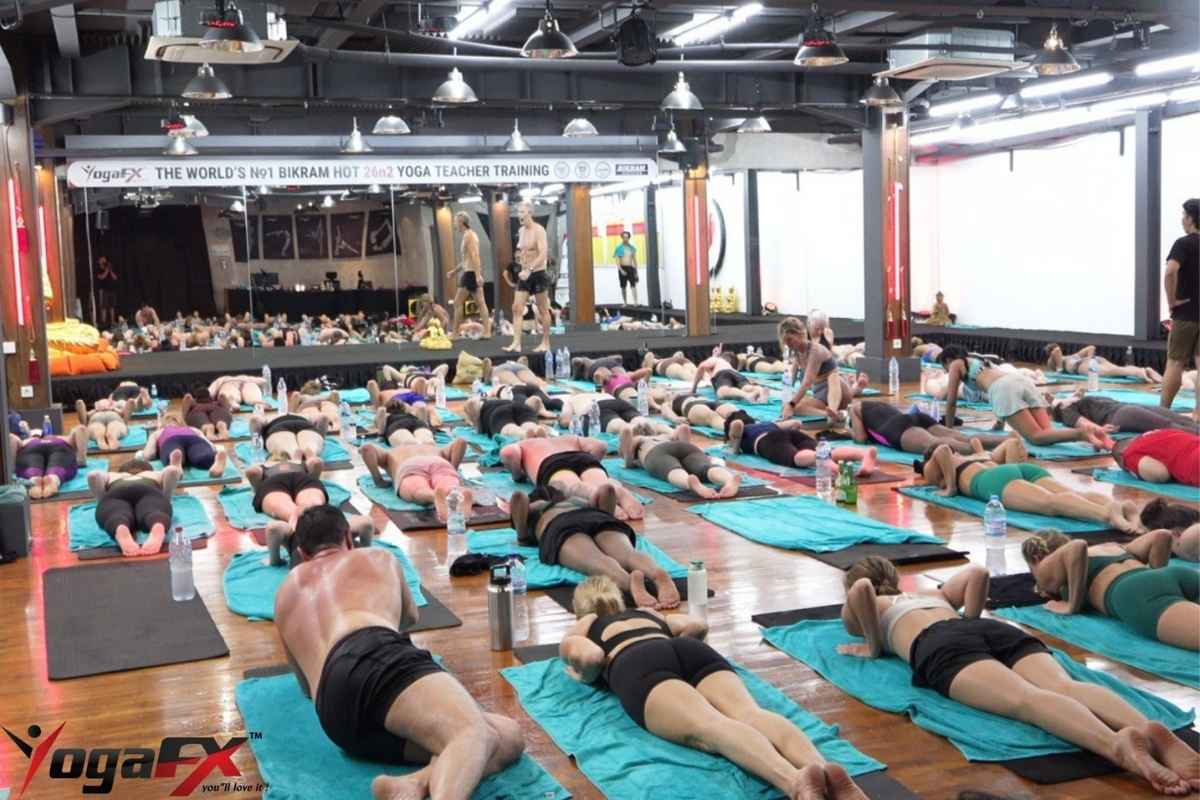 A full Bali hot yoga class lying face down on teal mat towels while the teacher walks the room under the YogaFX banner