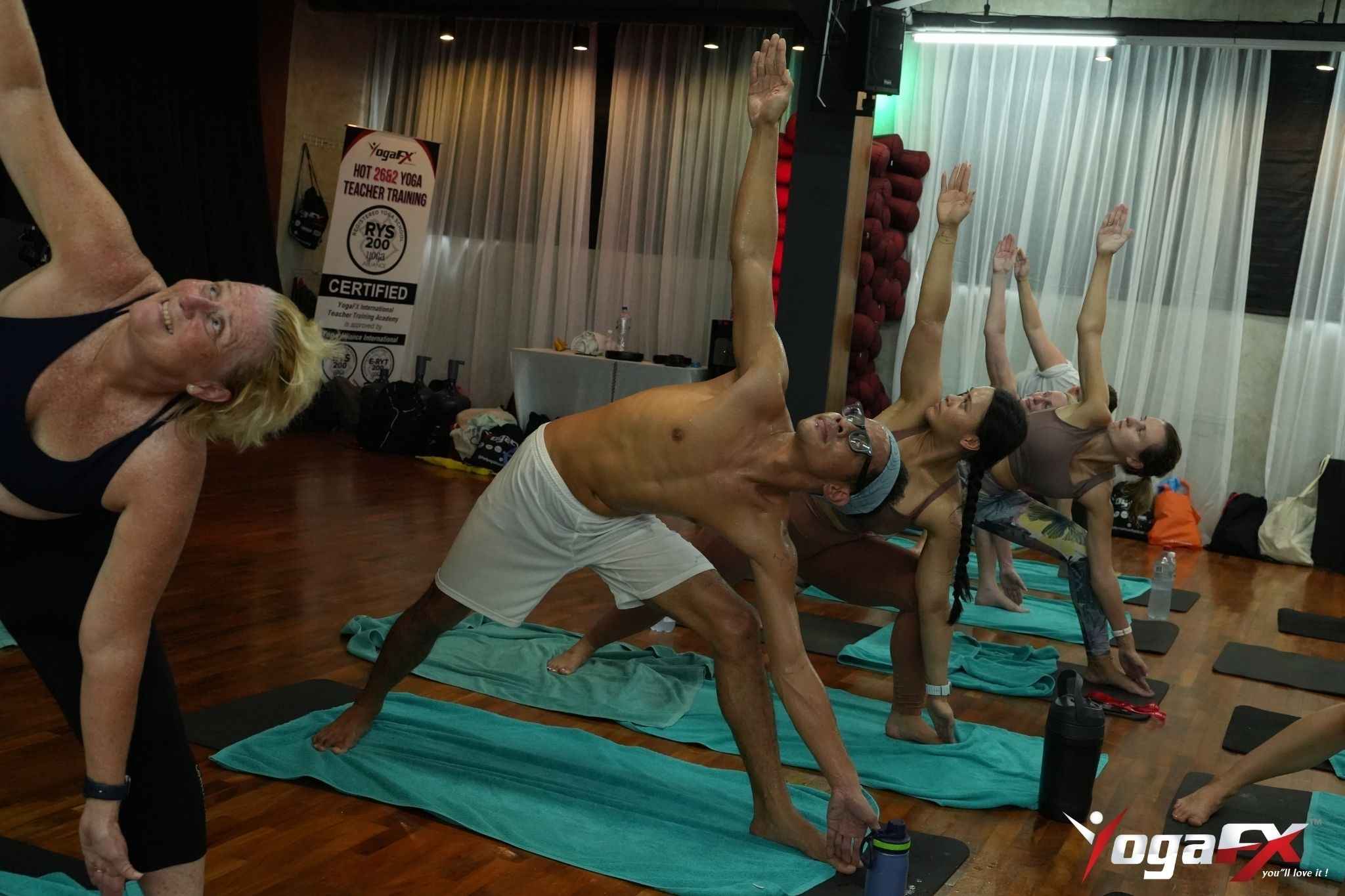 A man wearing a headband and white shorts in Triangle Pose, with a water bottle and towel on his mat in a Bali hot yoga class