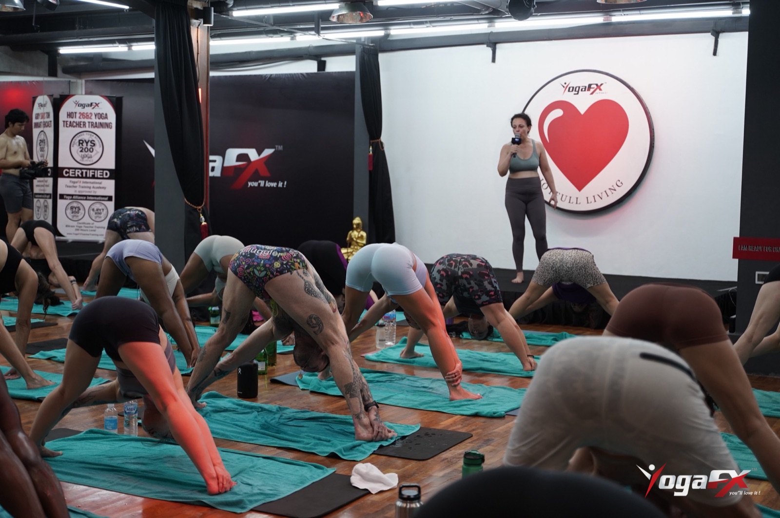 A full hot yoga class folding forward in Bali, with men in fitted shorts on teal towels in the front rows